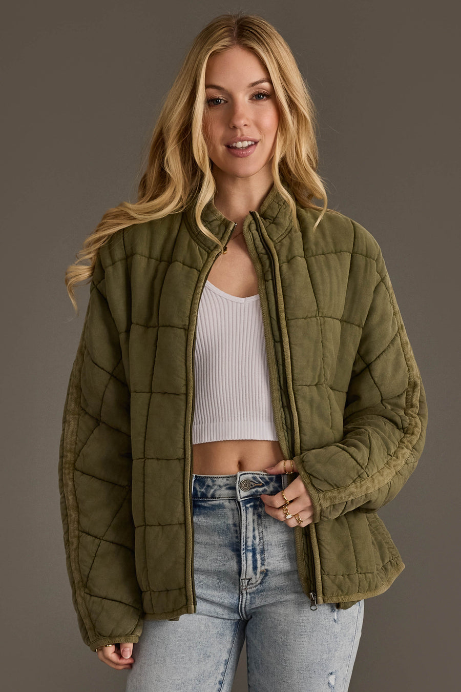 A woman smiles with one hand in her jeans pocket, wearing the Olive Soft Quilted Jacket from Panache Apparel Collection over a white ribbed crop top and light blue jeans, standing against a plain brown background.