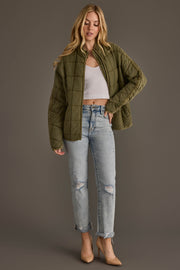 A woman wearing the Olive Soft Quilted Jacket from Panache Apparel Collection, paired with a white crop top, ripped light-wash jeans, and beige heels, stands against a plain gray background.