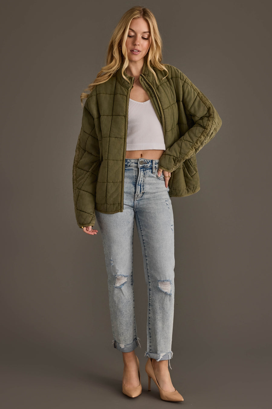 A woman wearing the Olive Soft Quilted Jacket from Panache Apparel Collection, paired with a white crop top, ripped light-wash jeans, and beige heels, stands against a plain gray background.