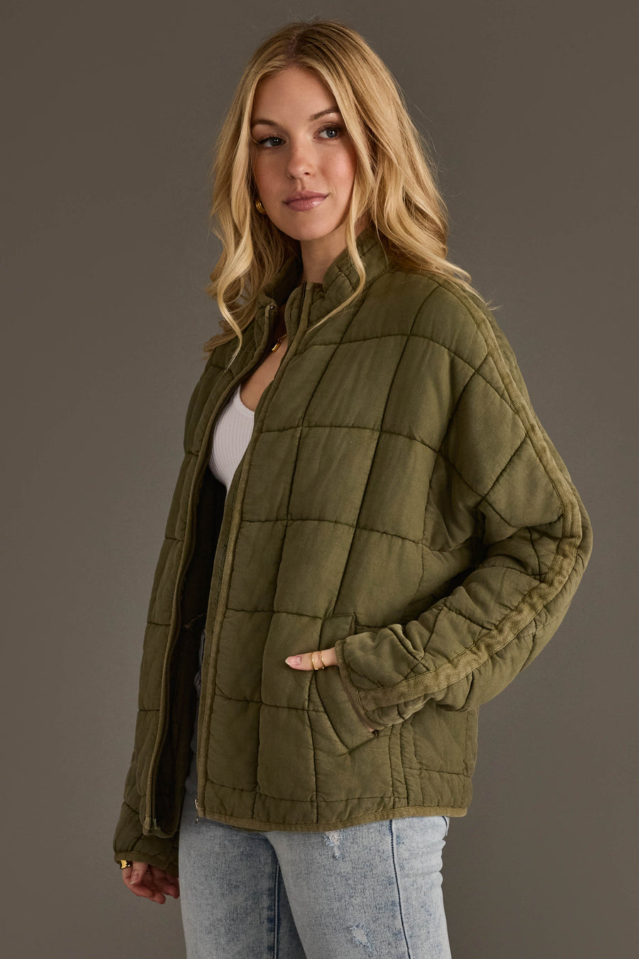 A woman models the Panache Apparel Collection’s Olive Soft Quilted Jacket, layered over a white top and paired with light blue jeans, standing with one hand in her pocket against a plain gray background.