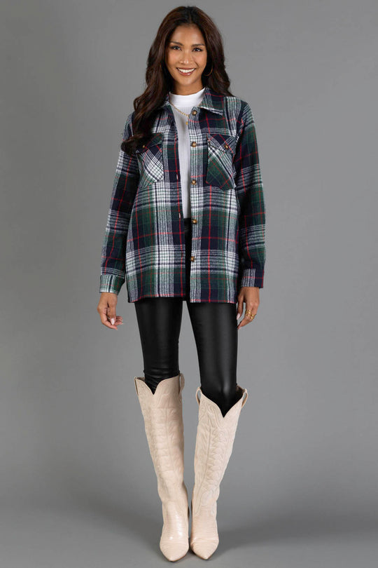 A woman stands smiling against a gray background, wearing the Panache Apparel Collection Navy, Green, White & Red Plaid Jacket, paired with black leather pants and tall cream cowboy boots.