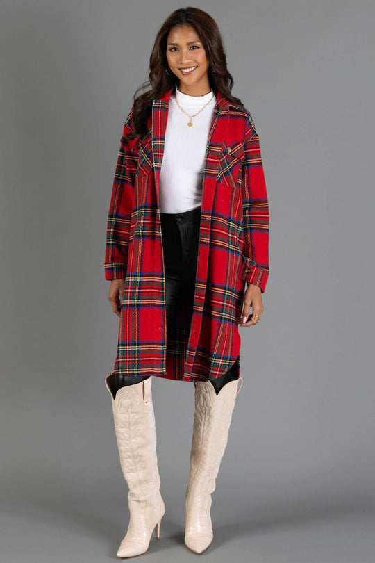 A woman stands and smiles against a plain gray background, wearing Panache Apparel Collection’s Red, Green, Blue & Yellow Plaid Long Shacket over a white turtleneck, paired with black pants and tall cream-colored cowboy boots.