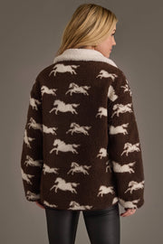 A person with long blonde hair is seen from the back wearing the Panache Apparel Collection Brown & White Fleece Horse Jacket, featuring a cozy brown fleece design with a white collar and distinctive white horse patterns. They also wear black pants and stand against a plain background.