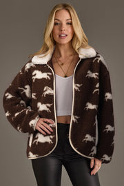 A woman models the Brown & White Fleece Horse Jacket from Panache Apparel Collection, featuring soft brown fleece adorned with white horse patterns, styled with a white crop top and black pants against a plain background.