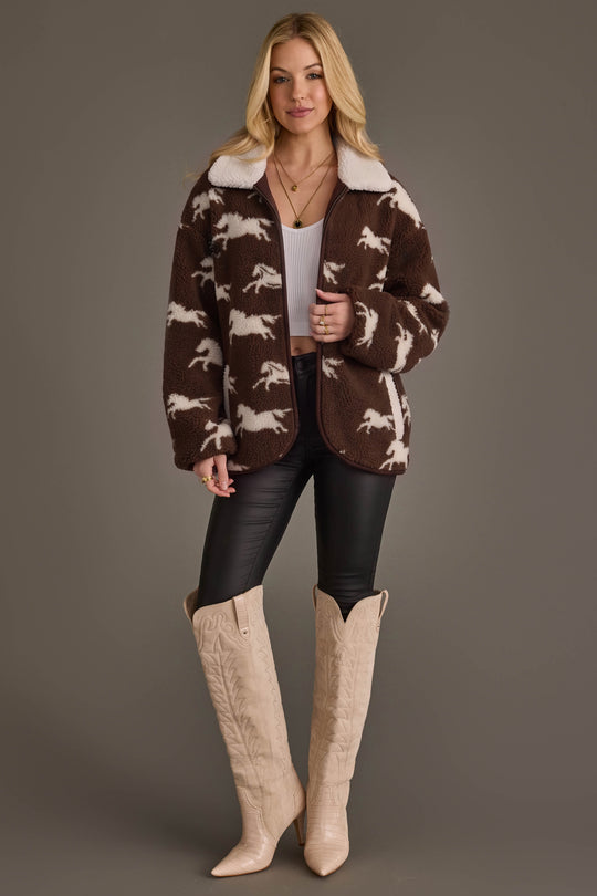 A woman stands against a plain background wearing the Panache Apparel Collection’s Brown & White Fleece Horse Jacket over a white top and black pants, paired with cream cowboy boots. She has long blonde hair and holds a cup in one hand.
