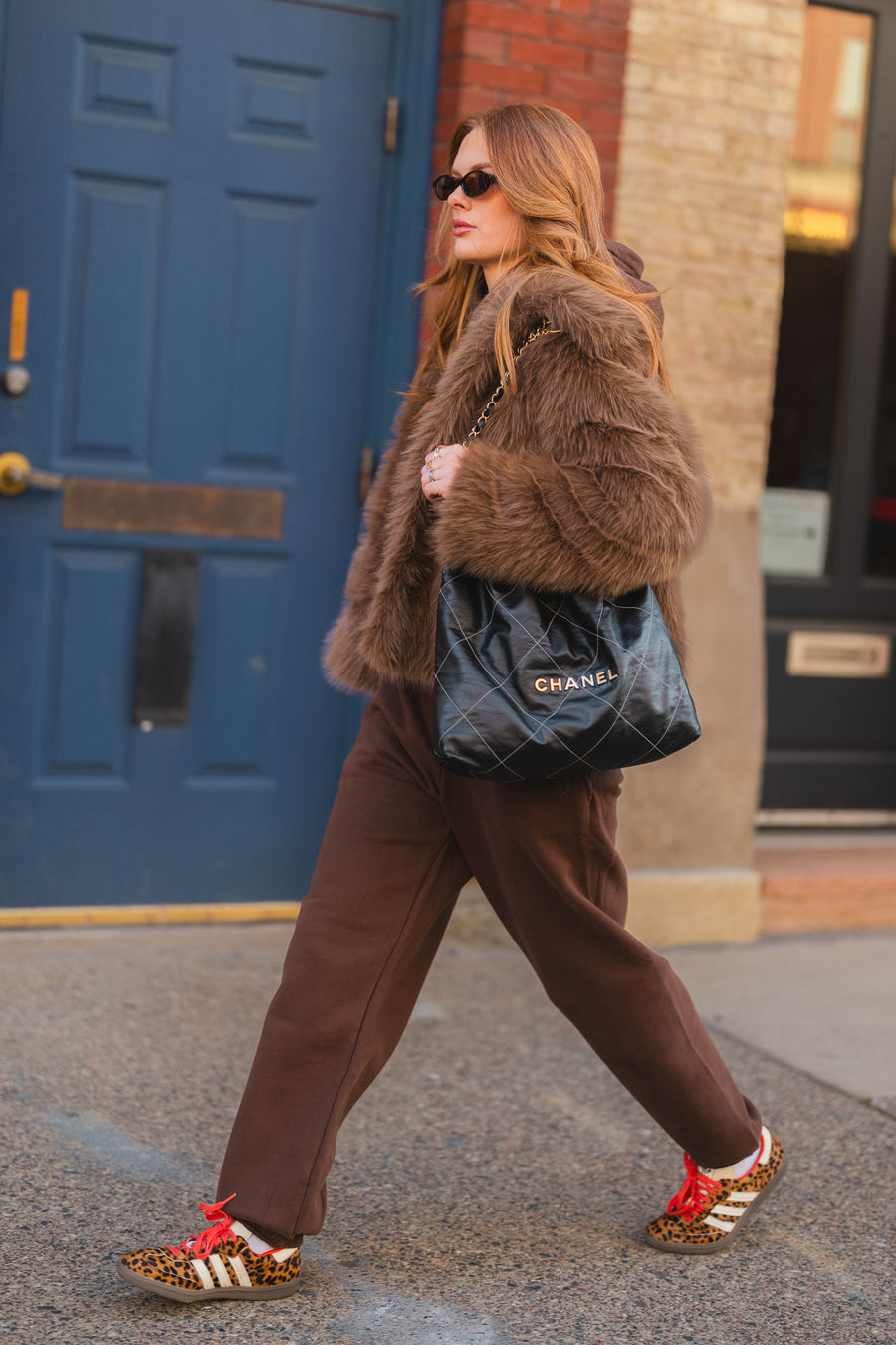 A person in a Light Brown Faux Fur Jacket by Panache Apparel Collection and brown sweatpants walks down a sidewalk carrying a black quilted handbag and wearing leopard print sneakers with red laces.