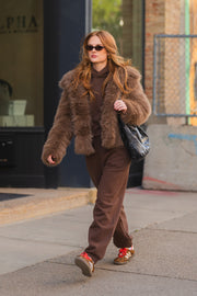 A person wearing a Light Brown Faux Fur Jacket, hoodie, and sweatpants walks down a sidewalk in sunglasses while carrying a black shoulder bag.