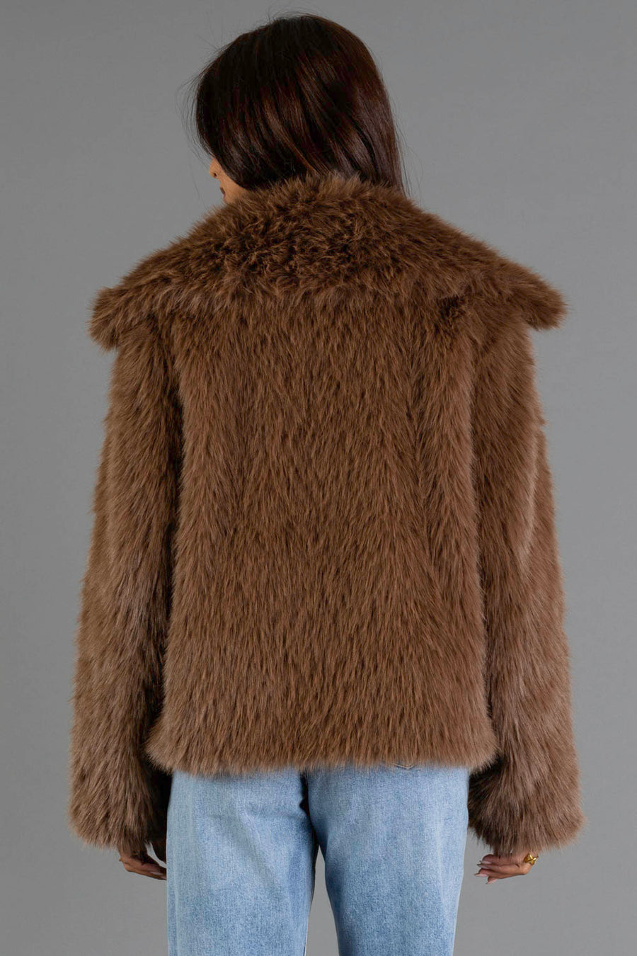 A person faces away from the camera wearing the Panache Apparel Collection Light Brown Faux Fur Jacket paired with light blue jeans, standing against a plain gray background.