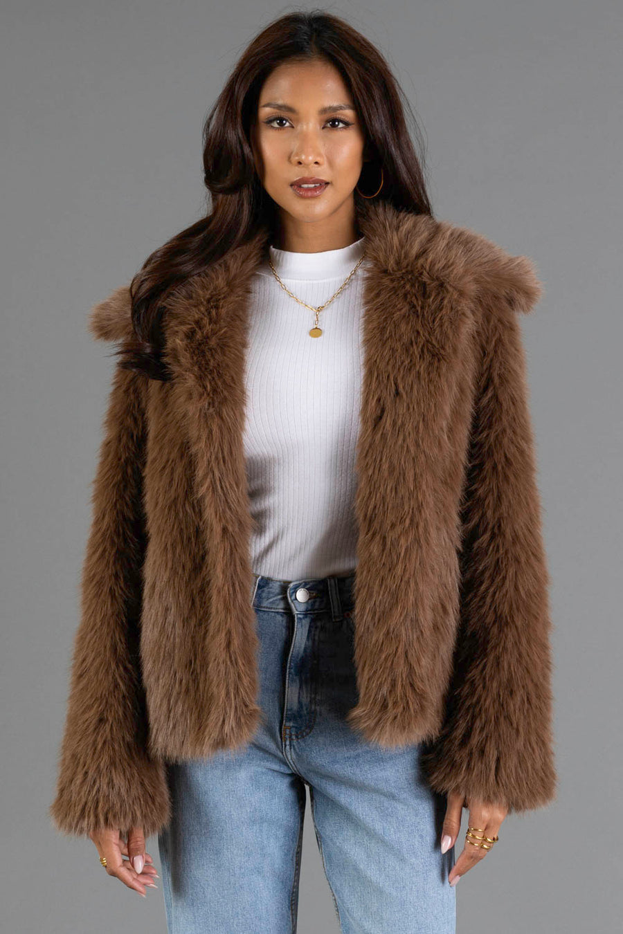 A woman models the Panache Apparel Collection Light Brown Faux Fur Jacket over a white ribbed turtleneck and blue jeans, standing against a plain gray background. She has long brown hair and wears gold hoop earrings.