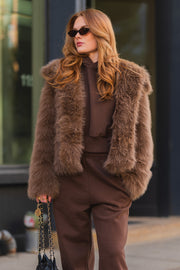A person wearing a Light Brown Faux Fur Jacket over a matching brown hooded sweatshirt and sweatants, carrying a small black bag with a gold chain strap.
