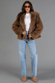 A woman stands confidently against a plain gray background, dressed in the Panache Apparel Collection Light Brown Faux Fur Jacket paired with a white turtleneck, light blue jeans, tan heels, and round sunglasses.