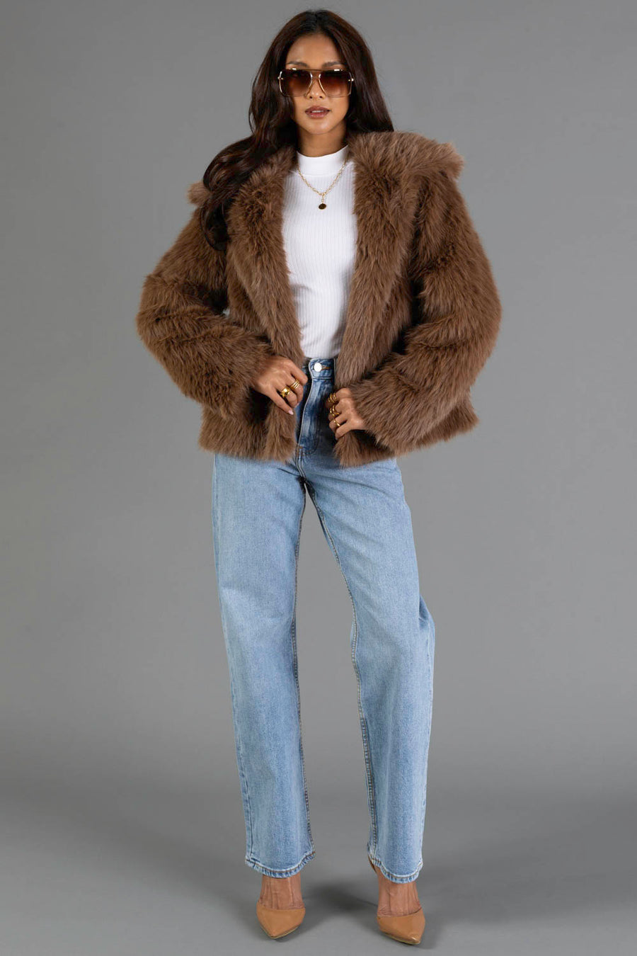 A woman stands confidently against a plain gray background, dressed in the Panache Apparel Collection Light Brown Faux Fur Jacket paired with a white turtleneck, light blue jeans, tan heels, and round sunglasses.