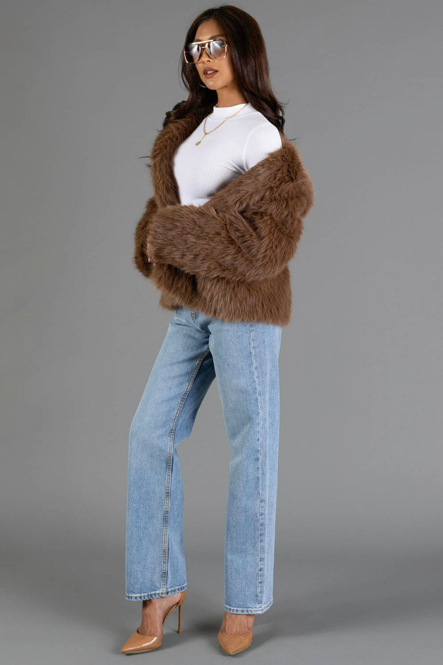 A woman in sunglasses wears a white top, the Light Brown Faux Fur Jacket from Panache Apparel Collection, light blue wide-leg jeans, and nude heels while standing against a gray background.