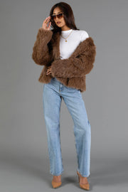 A woman wearing sunglasses, a white turtleneck, light blue jeans, nude heels, and the Light Brown Faux Fur Jacket from Panache Apparel Collection stands against a gray background, adjusting her sunglasses with one hand.