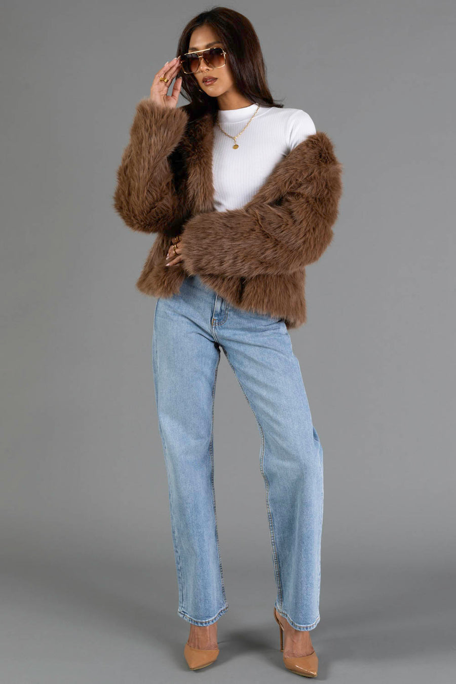 A woman wearing sunglasses, a white turtleneck, light blue jeans, nude heels, and the Light Brown Faux Fur Jacket from Panache Apparel Collection stands against a gray background, adjusting her sunglasses with one hand.