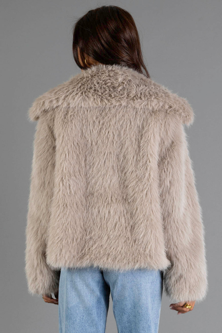 A person faces away from the camera, wearing the Panache Apparel Collection Taupe Faux Fur Jacket with light blue jeans against a plain gray background.
