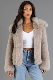 A woman wearing the Panache Apparel Collection Taupe Faux Fur Jacket over a white crop top and blue jeans, with long wavy hair, gold necklaces, and a neutral expression, stands against a plain gray background.