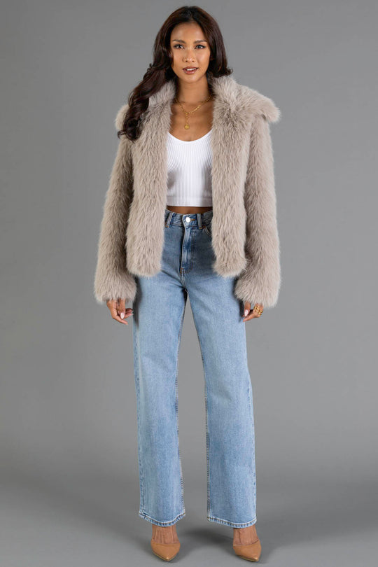 A woman with long, wavy hair and gold jewelry stands against a gray background wearing the Panache Apparel Collection’s Taupe Faux Fur Jacket, a white cropped tank top, light blue high-waisted jeans, and tan heels.