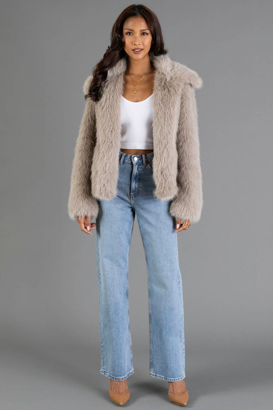 A woman with long, wavy hair and gold jewelry stands against a gray background wearing the Panache Apparel Collection’s Taupe Faux Fur Jacket, a white cropped tank top, light blue high-waisted jeans, and tan heels.