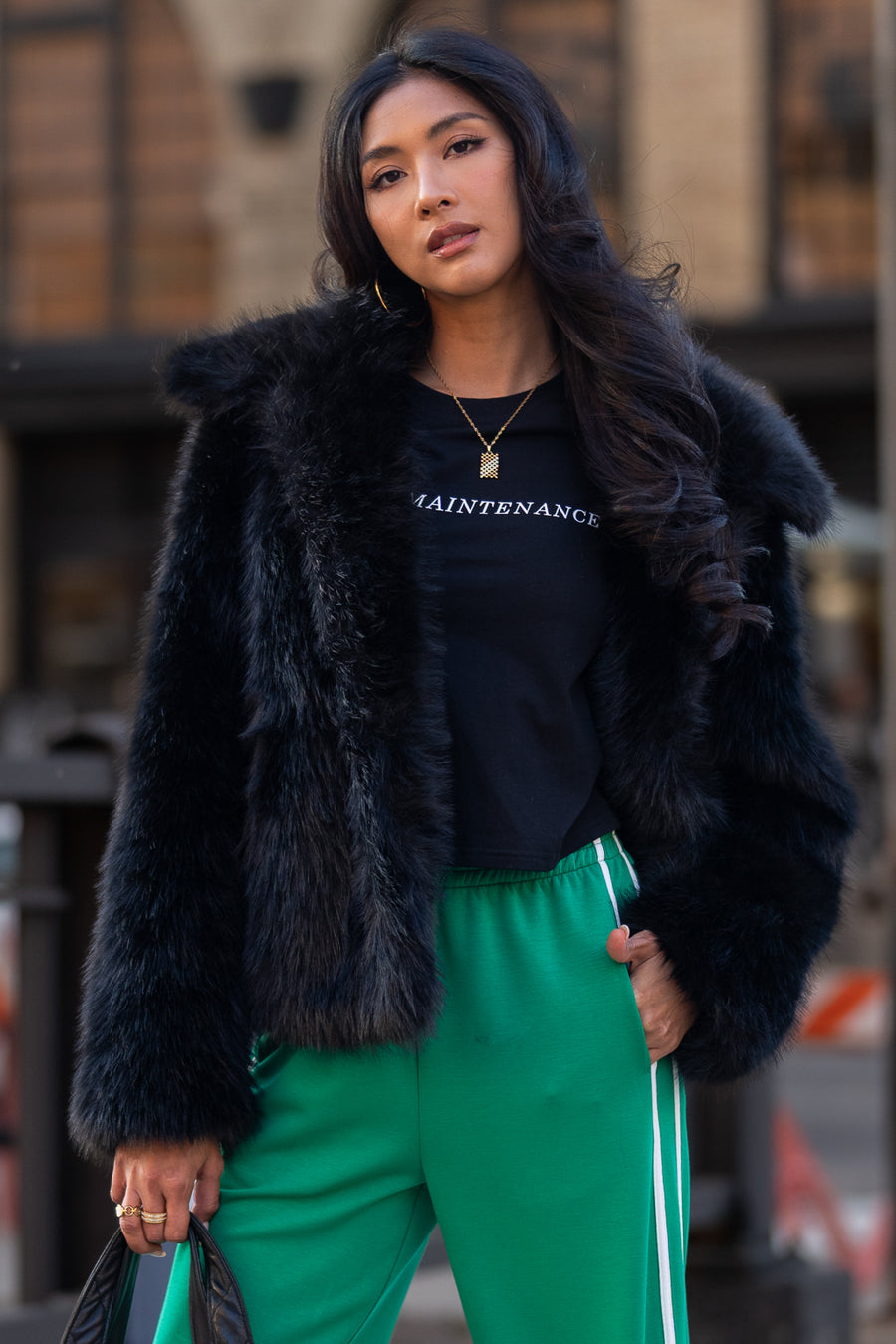A person wearing a Panache Apparel Collection Black Faux Fur Jacket over a black t-shirt and green sweatpants with white side stripes.