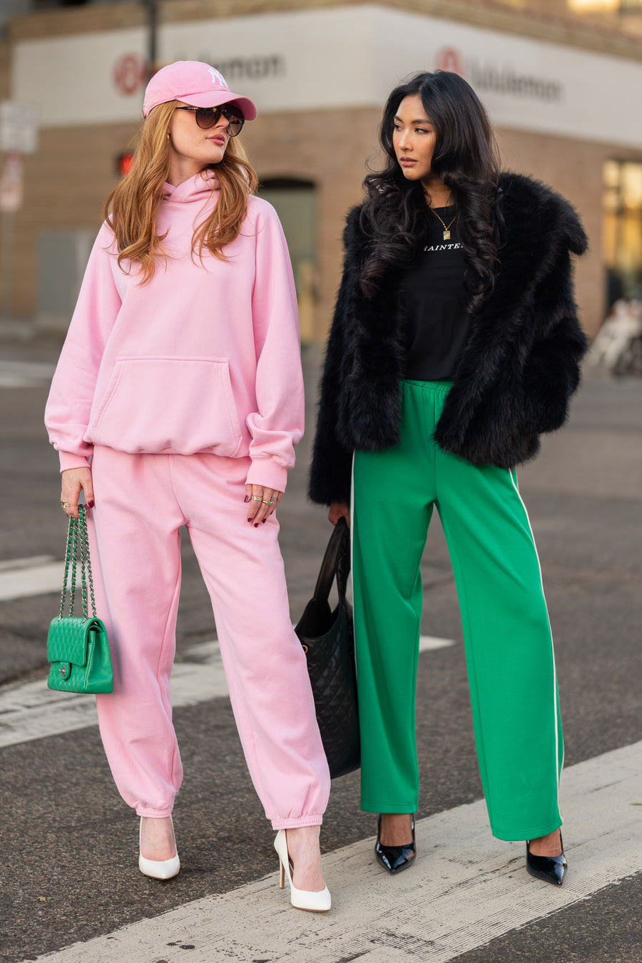Two women stand on a city street; one wears a pink hoodie and sweatpants with a green purse and white heels, while the other wears a Black Faux Fur Jacket by Panache Apparel Collection, black top, green pants, and black heels.