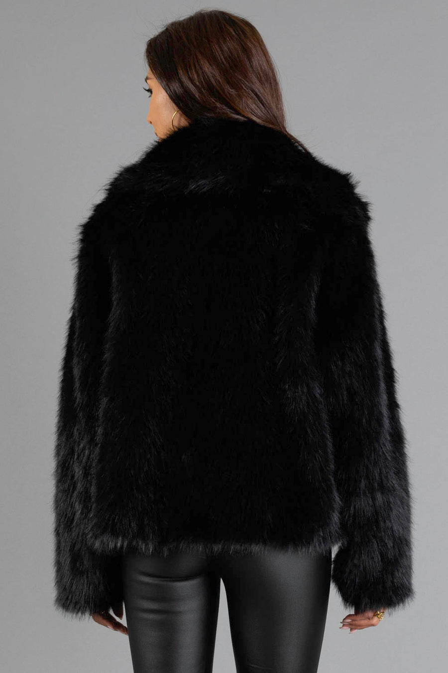 A person with long brown hair is shown from the back, standing against a plain gray background and wearing the Panache Apparel Collection Black Faux Fur Jacket paired with black leather pants.