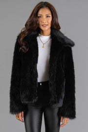 A woman is shown wearing the Panache Apparel Collection Black Faux Fur Jacket over a white ribbed top and black pants, accessorized with gold jewelry, standing against a plain gray background.
