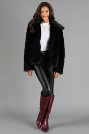 A woman stands and smiles against a plain gray background, wearing the Panache Apparel Collection Black Faux Fur Jacket, paired with a white turtleneck, black leather pants, and maroon knee-high boots.