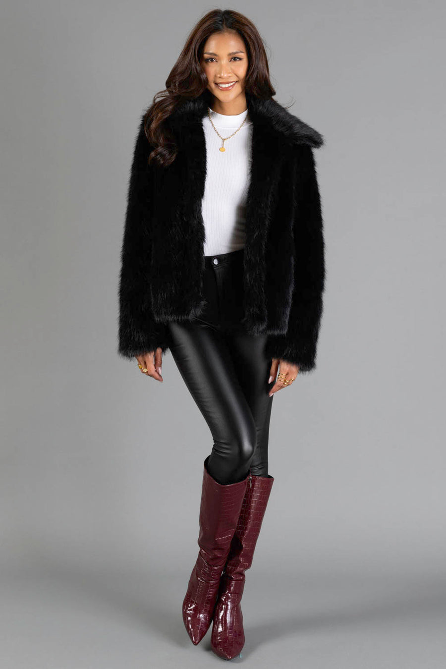 A woman stands and smiles against a plain gray background, wearing the Panache Apparel Collection Black Faux Fur Jacket, paired with a white turtleneck, black leather pants, and maroon knee-high boots.