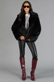 A woman wearing the Panache Apparel Collection Black Faux Fur Jacket, paired with black leggings, burgundy knee-high boots, sunglasses, and a white top, stands against a plain grey background.