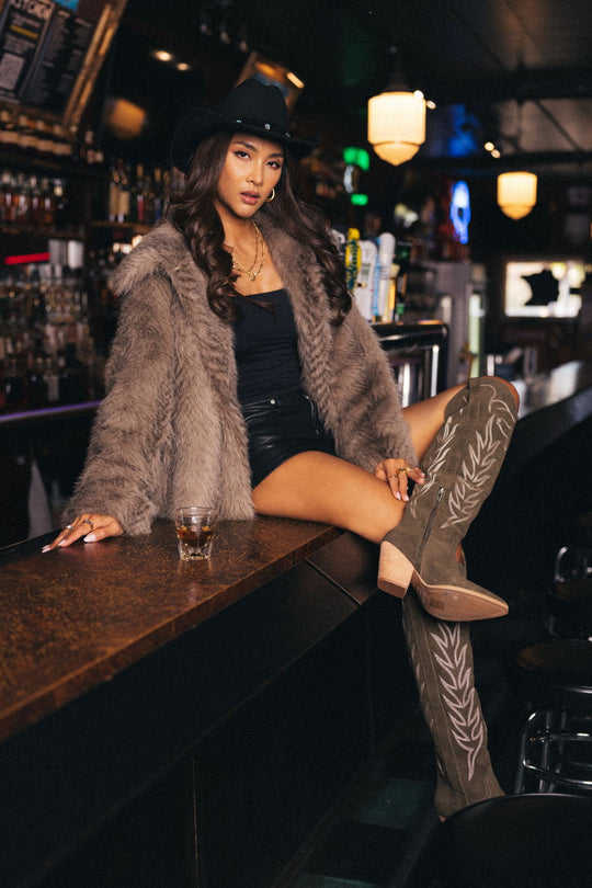 A woman wearing the Panache Apparel Collection Two Tone Faux Fur Jacket with an oversized collar, paired with a black hat, black top, and tall embroidered boots, sits on a bar counter in a dimly lit bar with a drink in front of her.