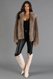A woman stands against a plain gray background wearing the Panache Apparel Collection Two Tone Faux Fur Jacket, paired with a black top, black leather pants, and cream knee-high boots.