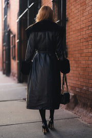 A person seen from behind wears a Black Faux Fur Trim Maxi Coat in black faux leather with a faux fur collar and cuffs, black tights, and high heels while carrying a small black chain-strap purse.
