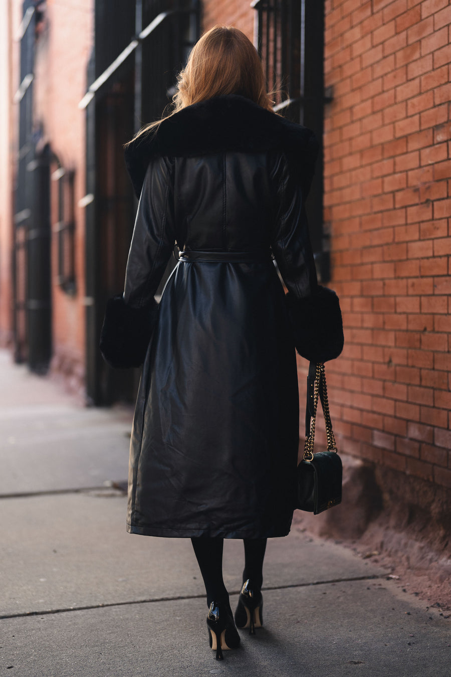 A person seen from behind wears a Black Faux Fur Trim Maxi Coat in black faux leather with a faux fur collar and cuffs, black tights, and high heels while carrying a small black chain-strap purse.