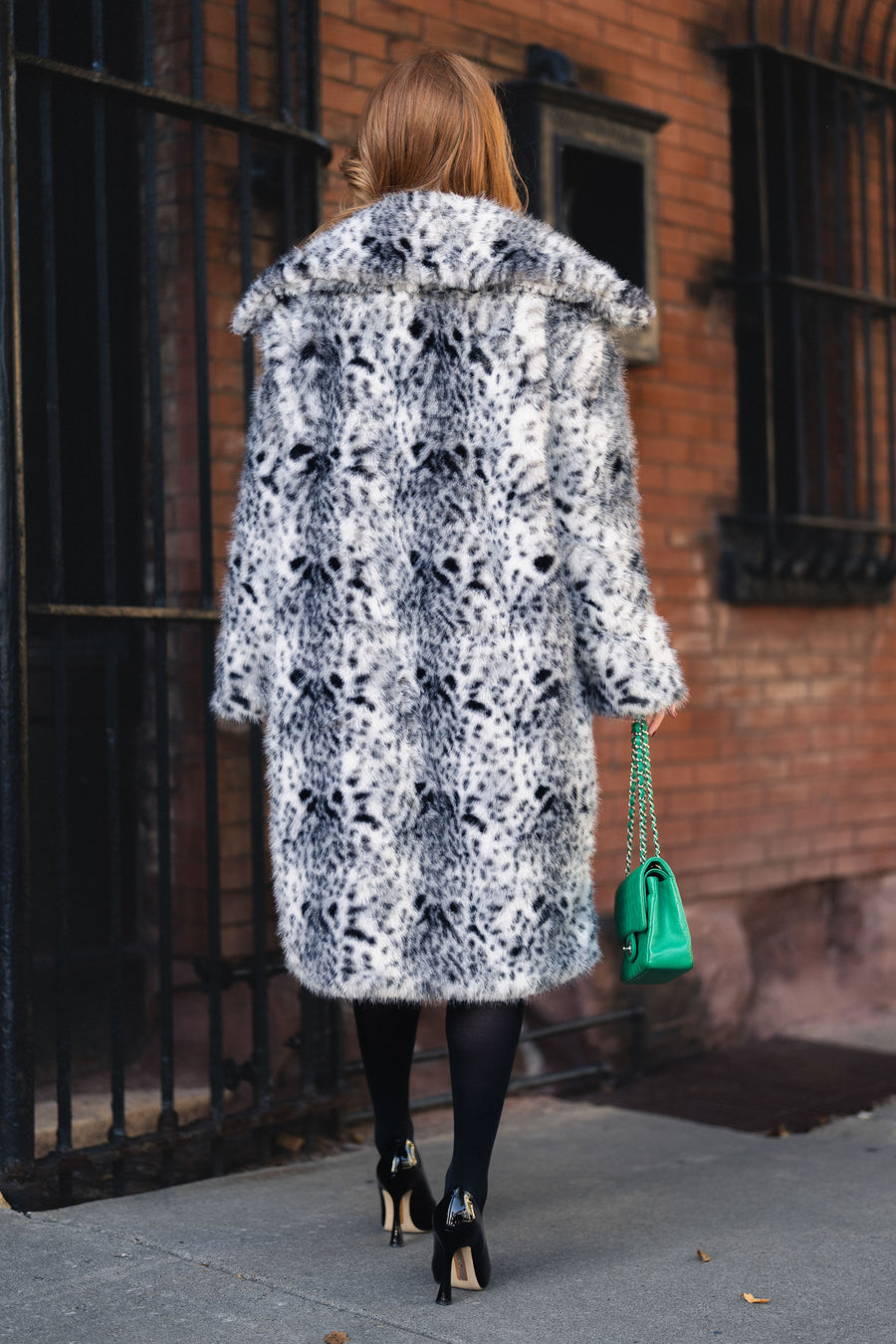 A person seen from behind wearing an oversized Leopard Faux Fur Maxi Coat from Panache Apparel Collection, black tights, black heels, and carrying a small green chain-strap handbag.