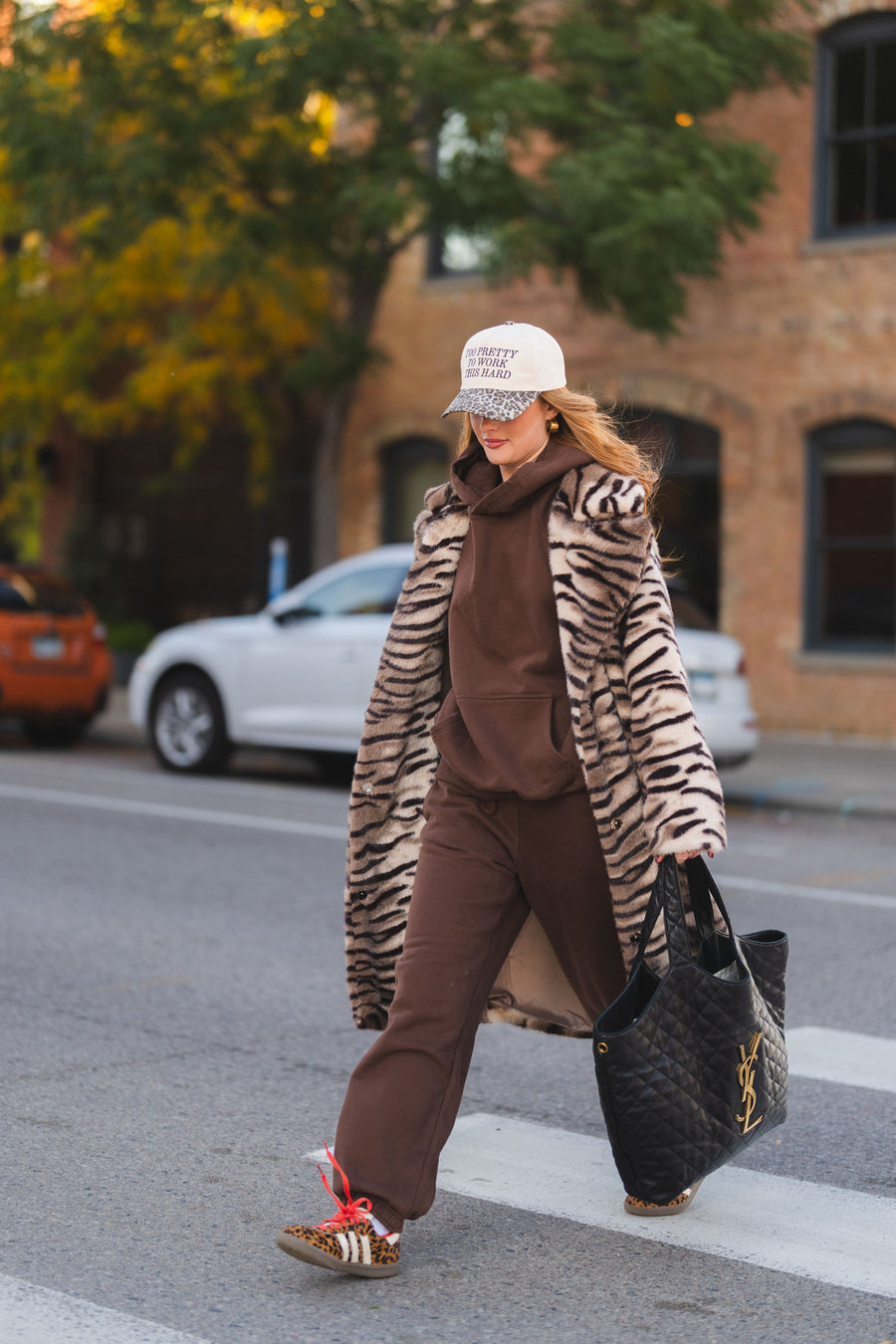 A person wearing a Panache Apparel Collection Tiger Stripe Faux Fur Maxi Coat over a brown hoodie and sweatpants, with a baseball cap, walks across a crosswalk carrying a large black quilted tote bag.