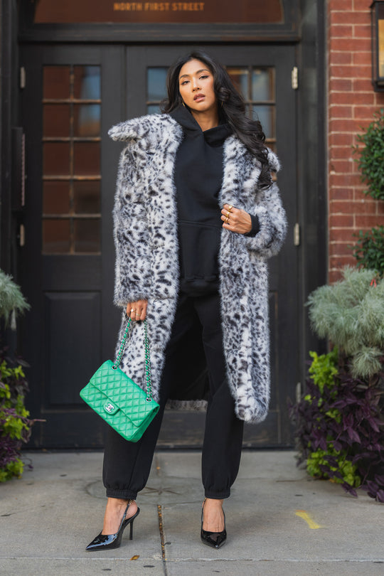 A woman wearing a Leopard Faux Fur Maxi Coat over a black hoodie and joggers, carrying a green quilted handbag and wearing black high-heeled pumps.