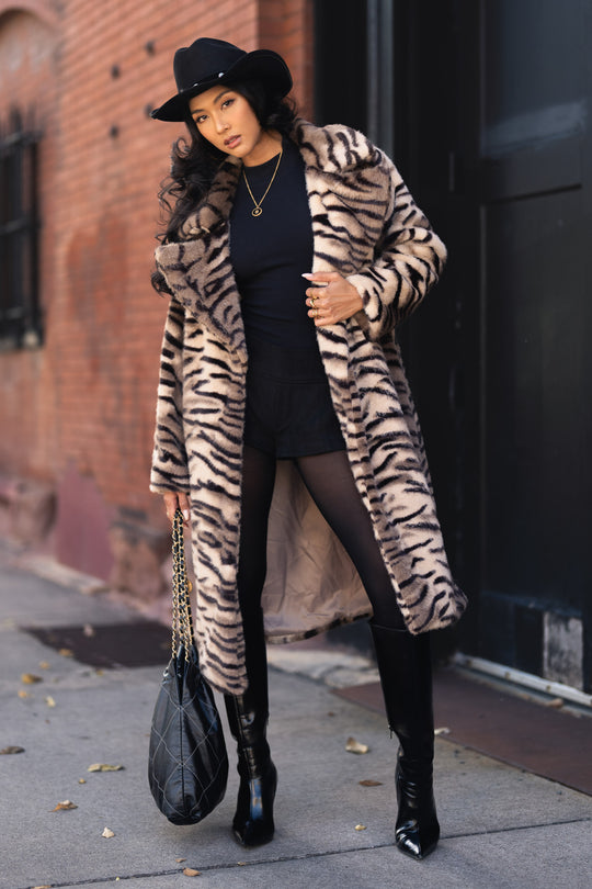 A woman wears a Panache Apparel Collection Tiger Stripe Faux Fur Maxi Coat, a fedora, black shorts, tights, and knee-high boots, while holding a quilted handbag.