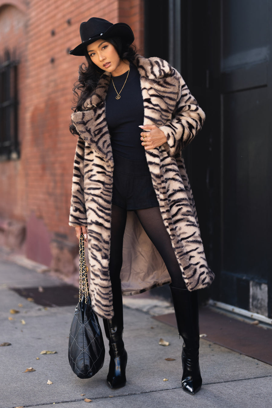 A woman wears a Panache Apparel Collection Tiger Stripe Faux Fur Maxi Coat, a fedora, black shorts, tights, and knee-high boots, while holding a quilted handbag.
