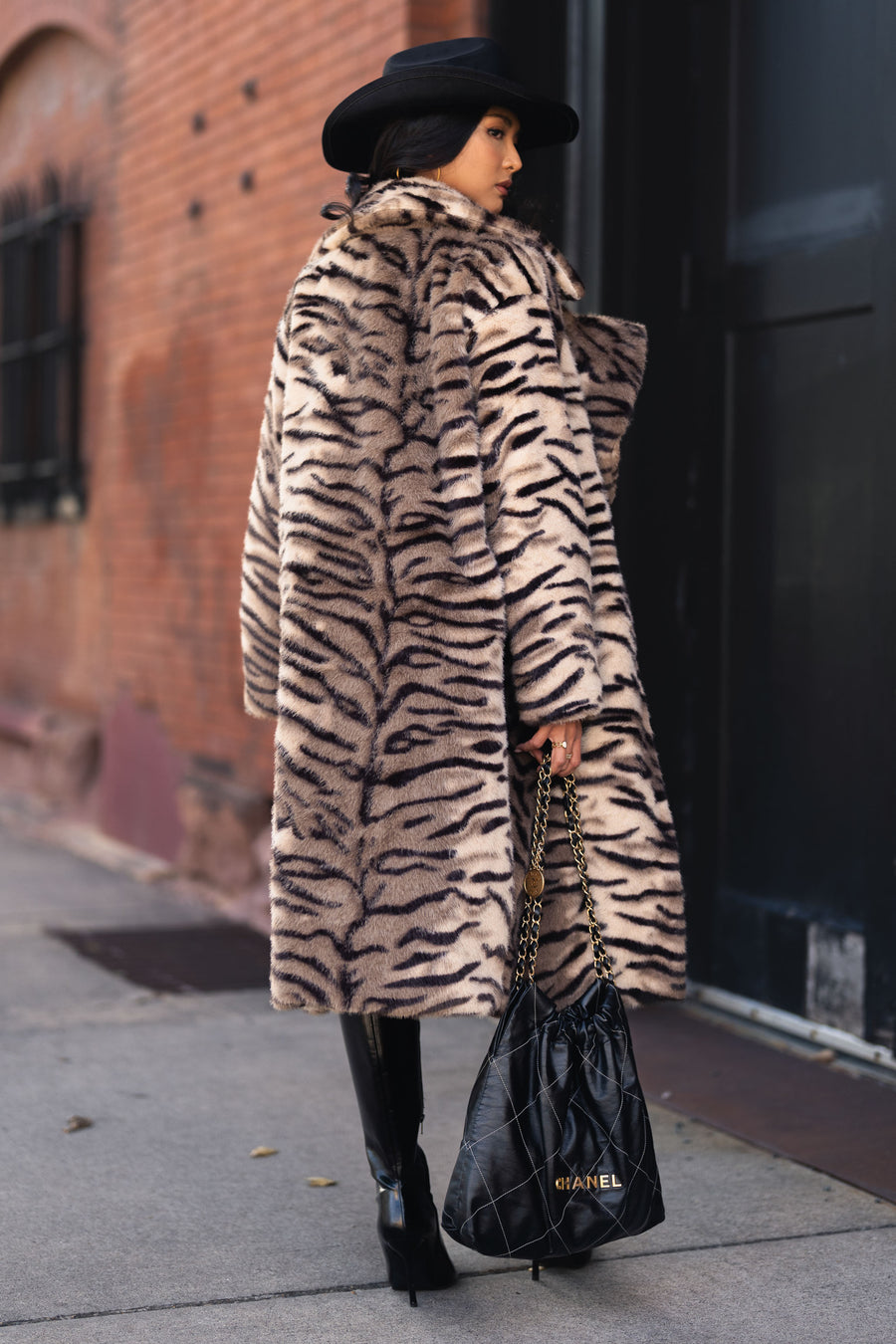A person wearing a Tiger Stripe Faux Fur Maxi Coat from Panache Apparel Collection, paired with a black hat, tall black boots, and a black quilted shoulder bag.