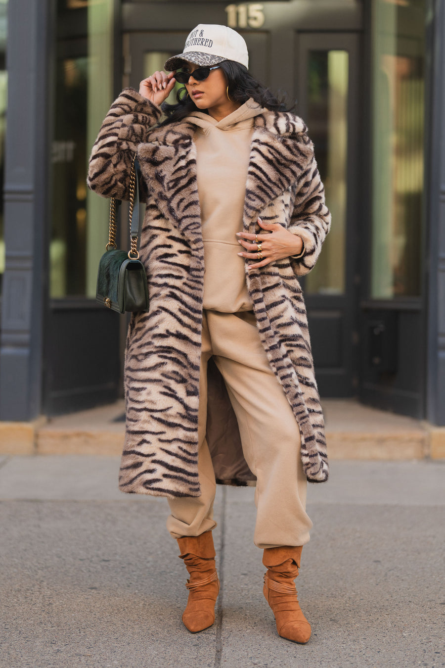 A person wearing a Panache Apparel Collection Tiger Stripe Faux Fur Maxi Coat over a beige hoodie and sweatpants, with a baseball cap, sunglasses, and tan slouchy boots.