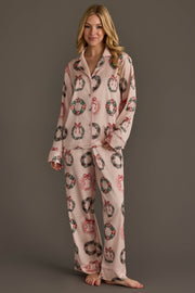 A woman wears a Christmas Wreaths Satin Pajama Set consisting of a long-sleeved, button-down pink top and matching pants featuring a pattern of wreaths with red bows.