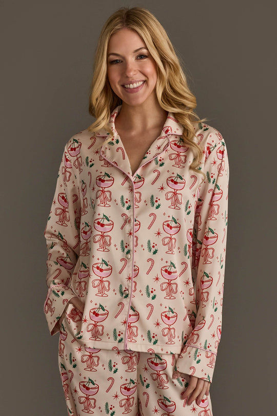 A woman wearing an Ivory Christmas Cocktails Satin Pajama Set, consisting of ivory long-sleeved pajamas with a festive cocktail print of glasses, cherries, candy canes, and holly leaves.