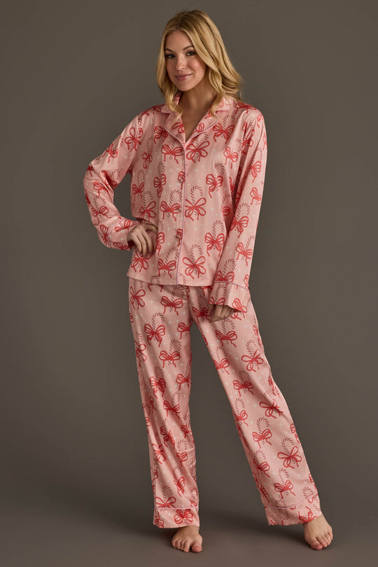 A woman models the Candy Canes & Bows Satin Pajama Set, a light pink button-down satin set featuring a red candy cane print.