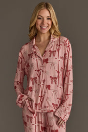 A woman wearing a light pink Bows Jersey Pajama Set by Panache Apparel Collection, featuring a long-sleeved button-up top and matching pants patterned with red bows.