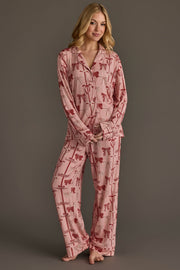 A woman wears a light pink Bows Jersey Pajama Set by Panache Apparel Collection, featuring a long-sleeved button-up top and matching pants patterned with festive dark red bows.