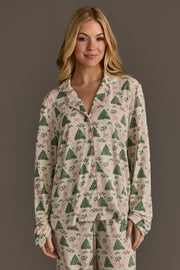 A woman wears the Panache Apparel Collection Trees & Bows Jersey Pajama Set, consisting of a long-sleeved button-down top and matching pants featuring a print of green Christmas trees and pink bows.