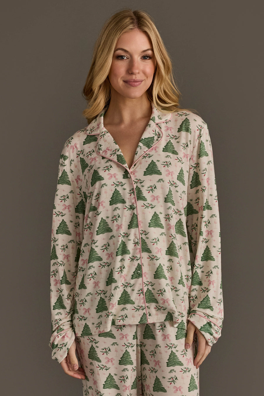 A woman wears the Panache Apparel Collection Trees & Bows Jersey Pajama Set, consisting of a long-sleeved button-down top and matching pants featuring a print of green Christmas trees and pink bows.