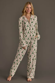 A woman wearing the Panache Apparel Collection Trees & Bows Jersey Pajama Set, featuring a long-sleeved button-up top and matching pants decorated with a pattern of green Christmas trees and pink bows.