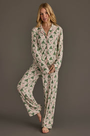 A woman wears a Panache Apparel Collection Trees & Bows Jersey Pajama Set consisting of a long-sleeved button-down top and matching pants patterned with green pine trees and pink bows.